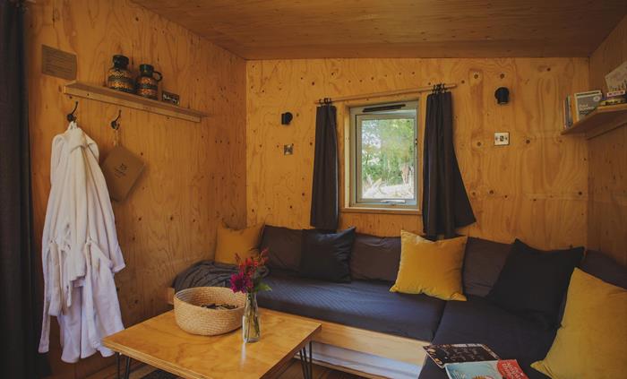 an indoor scene of a cabin with a large corner sofa strewn with scatter cushions and a magazine, a coffee table on a rug sits in the foreground. there